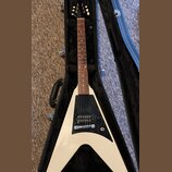 Bílá elektrická kytara s trojúhelníkovým tělem v černém kufru. Nástroj má hnědý hmatník, tři snímače a černý pickguard. Na hlavě kytary není žádný vi…