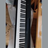 Černý digitální klavír s bílými a černými klávesami. Na horním panelu je uprostřed bílé barvy nápis YAMAHA s doprovodným logem. V levé části panelu j…