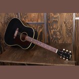 Černá akustická kytara s hnědým rýhovaným plechtrem a bílým lemováním korpusu. Na hlavě nástroje je zlatý nápis Gibson. Hриф má tmavou hmatovnici s j…
