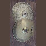 Dvě kovové činely položené na hnědém laminátovém povrchu. Spodní činel má černý nápis PAiste 302 a pod ním menší text 14" HI-HAT Bottom. Horní činel…