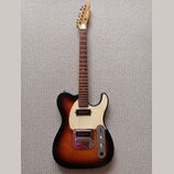 Elektrická kytara typu Telecaster s tělem v provedení sunburst a bílým pickguardem. Na hlavě hředu je vidět nápis "Aria". Kytara má dva snímače, kovo…