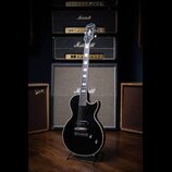 Černá elektrická kytara s tmelem hmatníkem a chromovaným hardwarem, umístěná na stojanu. Na hlavě hmatníku je bílé logo Gretsch a na korpusu pod sním…