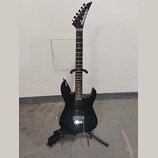 Černá elektrická kytara s špičatou hlavou, na které je bílé logo Jackson. Nástroj má tmavý hmatník a stříbrný kobylku typu Floyd Rose. Kytara je umís…