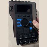 Černý elektronický přístroj s displejem a ovládacími prvky. Na levé straně je bílé logo s písmenem B a nápisem BEAT. Na spodní části jsou tlačítka s…