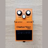 Oranžový kytarový efekt s nápisy Distortion a DS-1. Na horní části jsou tři ovládací knoflíky s popisky TONE, LEVEL a DIST oraz kontrolka CHECK. Ve s…