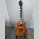 Elektrická kytara s trupem v žlutě-oranžovém sunburst provedení a hnědým hmatníkem s trapezoidními vložkami. Na hlavě je bílý nápis Gibson a kurzivní…