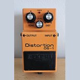 Oranžový kytarový efekt s nápisy Distortion a DS-1. Na horní části jsou tři ovládací knoflíky s popisky TONE, LEVEL a DIST a červená kontrolka CHECK.…