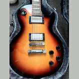 Elektrická kytara s tmelem v odstínu sunburst, umístěná v otevřeném pouzdře s šedým polstrováním. Nástroj má dva humbuckery s bílými kryty, chromovan…