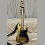 Zlatá elektrická basová kytara s černým pickguardem a javorovým hnedým hmatníkem. Na hlavě nástroje je nápis Fender Precision Bass. Kytara má čtyři s…