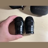 Dvě černé fürheadové kapsle s mřížkou. Na obou je vidět nápis SHURE, na pravé kapsli je dále uveden model NXN8/C. V pozadí je černý obal s nápisem SH…