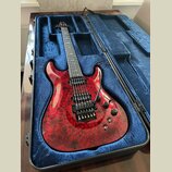 Elektrická kytara s červeným mramorovaným povrchem těla v černém tvrdém kufru s modrou výstuhou. Nástroj má šest strun, dva humbuckery a dva ovládací…