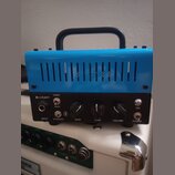 Modrý přenosný zesilovač s černým ovládacím panelem a černou rukojetí. Na panelu je nápis BLUEJAY, vstup INPUT a ovládací prvky s popisky BLUETOOTH (…