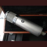 Stříbrný dynamický mikrofon s kovovou síťkou v dřevěném kufru s černým polstrováním. Na těle mikrofonu je umístěno kulaté logo s písmeny „us“. Povrch…