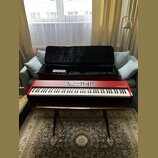 Červený digitální klavír s bílými a černými klávesami. Na pravé straně korpusu je bílý nápis nord piano. Nástroj leží na hnědém dřevěném stole před o…