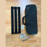 Na fotografii je černý kovový pedalboard s třemi vodicími lištami, černý přepravní vak s rukojetí a rolka kabelu v plastovém sáčku. Na vaku je modrý…