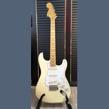 Krémově bílá elektrická kytara s jasným javorovým hnedlem krku a hmatníkem. Na hlavě je viditelné logo Fender. Kytara má tři snímače, bílý pickguard…