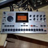 Stříbrný elektronický bicí automat s nápisem MACHINEDRUM na displeji a logem ELEKTRON v pravém horním rohu. Zařízení má digitální displej, několik ot…