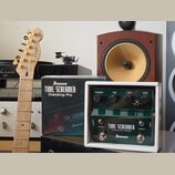 Na fotografii je zelený kytarový efekt v bílém ochranném boxu s nápisy TUBE SCREAMER, Overdrive Pro TS808DX a značkou Ibanez. Vedle efektu je umístěn…