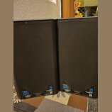 Dvě černé reproduktorové bedny s kovovou mřížkou. Na spodní části jsou viditelné nápisy K12.2, 2000 WATT a logo QSC s označením K.2 SERIES. Dále je z…