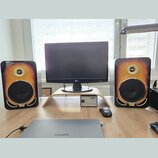 Dva studiové monitory s dřevěným provedením v přechodové barvě od žluté po tmavě hnědou. Na spodní části každého reproduktoru je viditelné logo Gibso…