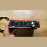 Červený audio rozhraní s černým předním panelem. Na zařízení jsou nápisy Scarlett 2i2, LINE INST, GAIN, DIRECT MONITOR, OFF ON, 48V a MONITOR. Jsou z…