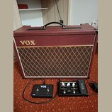 Vínový kytarový kombozesilovač s logem VOX a mřížkou v kosočtvercovém vzoru. Před zesilovačem jsou umístěny dvě černá pedály; levý nese nápis VOX a V…