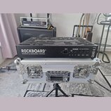 Černý pedalboard RockBoard QUAD 4.1 by WARWICK umístěný na stříbrném transportním kufru s kovovými hranami a zámky. Na boku pedalboardu je vidět pane…