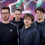 Na fotografii jsou čtyři muži v tmavém oblečení před barevnou zdí s graffiti. Jeden z nich má na černé mikině bílé logo adidas. Žádné hudební nástroj…