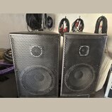 Dva černé reproduktorové boxy s kovovými perforovanými mřížkami. Každý box obsahuje jeden velký nízkofrekvenční a jeden malý vysokofrekvenční reprodu…