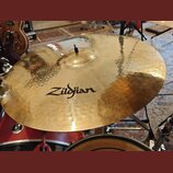 Zlatavý kovový činel s viditelným kovánívým povrchem a mírným opotřebením. Uprostřed je černý nápis Zildjian. Činel je osazen na stojanu v rámci bicí…