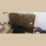 Hnědé pianino s vysokým leskem a dřevěnou strukturou. Před nástrojem stojí černá pianistická židlička s čalouněným sedadlem. Na nástroji ani v jeho o…