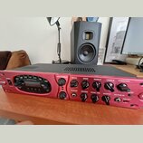 Červený kytarový zesilovač s černým horním krytem. Na předním panelu jsou nápisy POWER, AMP MODELS, DRIVE, BASS, MIDDLE, TREBLE a PRESENCE. V levé čá…