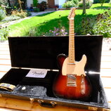 Elektrická kytara s tmeleým sunburst povrchem těla a světlým hmatníkem z javoru. Nástroj má bílý pickguard a na hlavě je viditelné logo Fender. Kytar…