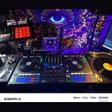 Fotografie zachycuje DJ set umístěný před barevným gobelínem s motivem oka. Uprostřed je černý DJ kontrolér se značkami RANE a serato, vybavený dvěma…