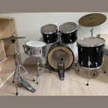 Sada akustických bubnů v černé barvě s kovovými obručmi. Obsahuje basový buben, dva tom-tomy, floor tom a stříbrný snare drum na stojanu. Na jednom z…