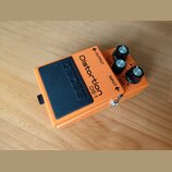 Oranžový efekt pedál s černým gumovým spínacím tlačítkem, na kterém je vyražený nápis BOSS. Na horní části je uveden název Distortion DS-1 a popisky…