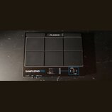 Černý elektronický sampling pad s šesti gumovými polštářky v provedení 2x3. V horní části je logo ALESIS a v dolní části nápis SAMPLEPAD PRO. V pravé…