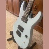 Světle modrá elektrická kytara s tmavým hnedým hmatníkem. Nástroj má tři snímače typu humbucker a jeden ovládací knoflík s nápisem TONE. Povrch kytar…