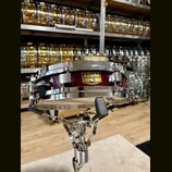 Vínový malý buben s chromovaným obručováním a mechanismy, umístěný na chromovaném stojanu. Na korpusu je zlatý štítek s nápisy TAMA, SNARE DRUM a Ser…