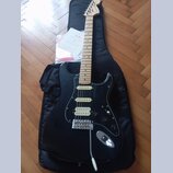 Černá elektrická kytara typu Stratocaster s bílým pickguardem a světlou hnedou hmatníkem. Na hlavě kytary je viditelné logo Fender a v pozadí leží pa…