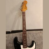 Černá elektrická kytara s hnědým hmatníkem a bílým pickguardem. Na hlavě nástroje je vidět logo Fender® STRATOCASTER. Povrch kytary vykazuje známky o…
