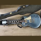 Modrá elektrická kytara v černém tvrdém kufru. Na hlavě je viditelné logo Epiphone. Kytara má dva humbuckery, bílý pickguard a čtyři ovládací knoflík…