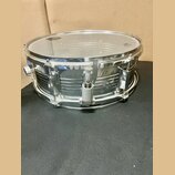 Stříbrný kovový snare drum s průhlednou blánou. Nástroj nemá žádné viditelné značky ani nápisy. Povrch kovu vykazuje drobné škrábance a známky opotře…