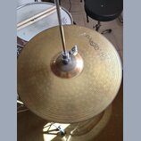 Zlatý kovový činel s viditelným rýhováním povrchu, namontovaný na kovovém stojanu. Na povrchu je čitelně napsán nápis Paiste 2002. V pozadí je vidět…
