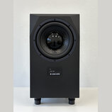 Černý studiový subwoofer v hranatém korpusu s matným povrchem. Na přední straně je umístěn velký kruhový reproduktor a pod ním ovládací panel s nápis…