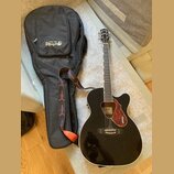 Černá akustická kytara s vyřezem v těle a hnědým rýhovaným plektrem. Na hlavě nástroje je nápis GRETSCH a na plektru jsou dvě loga GRETSCH. Kytara je…
