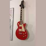 Červená elektrická kytara s tvarem single-cut, dvěma humbuckery a bílým pickguardem. Na hlavě hmatníku je viditelné logo "Kramer". Nástroj visí na ná…