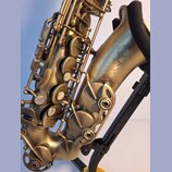 Detail saxofonu v matné zlaté barvě, který je umístěn na černém stojanu. Na korpu nástroje je vyryto logo SELMER PARIS a nápis MADE IN FRANCE. Pod ní…