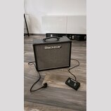 Černý kytarový kombo zesilovač s šedým textilním krytem reproduktoru a nápisem Blackstar. K zesilovači je připojen černý nožní spínač s logem Blackst…
