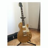 Zlatá elektrická kytara s tvarem singlecut, bílým pickguardem a dvěma humbuckery. Na hlavě hředu je viditelné logo Epiphone. Nástroj je umístěn na če…